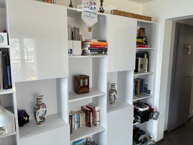 A well-organized bookshelf filled with books, decorative vases, a small toy, and tools, with a cabinet and a door visible in the background.