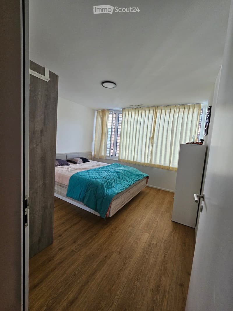 A well-lit bedroom with wooden flooring, a bed with a teal quilt and pillows, a wardrobe, and a window with curtains.