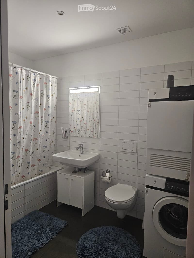 Modern bathroom with white tiles, a sink, toilet, bathtub, and a washing machine. Features include a floral shower curtain, a mirror, and two rugs on the floor.