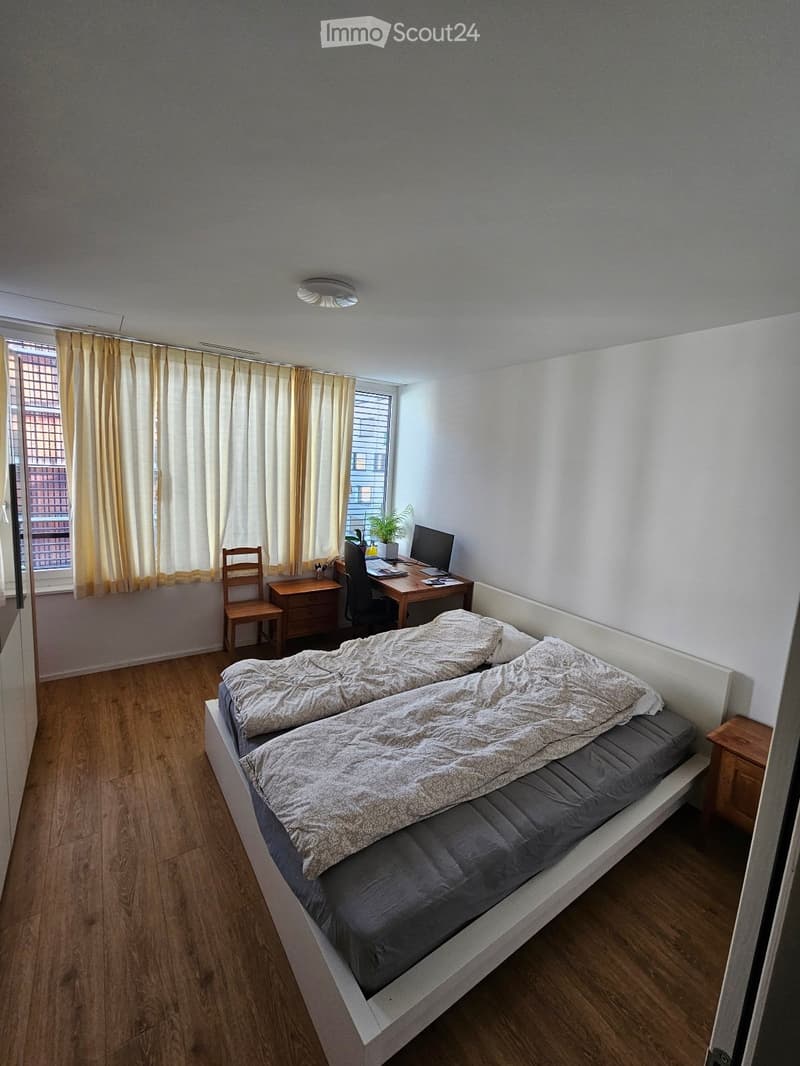 A tidy bedroom with a double bed, a wooden desk with a laptop, a chair, and a small plant near the window. The room features light-colored curtains and a wooden floor.