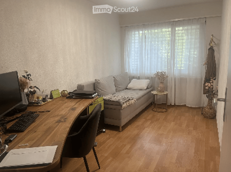 A small, well-lit living room with a grey sofa, a wooden desk with a laptop, a chair, and a small side table with a vase. The room has wooden flooring and large windows with white curtains.