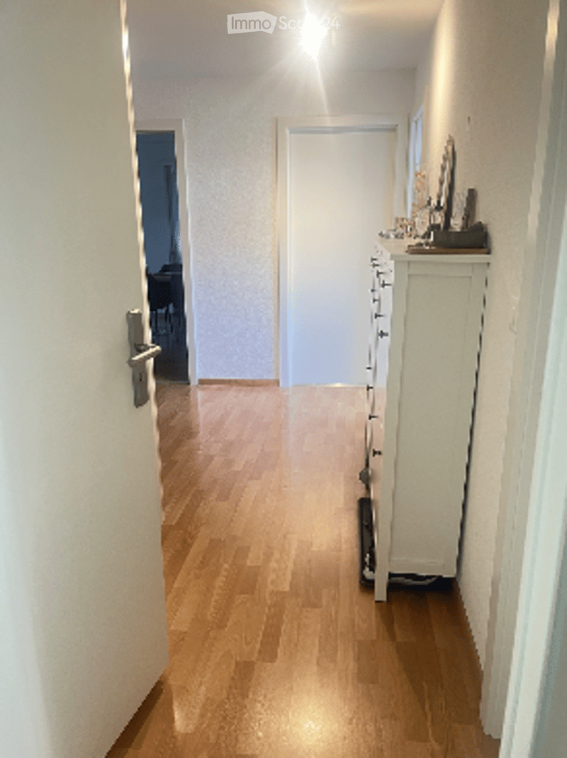 A narrow hallway with light wooden flooring, white walls, and a closed door at the end. A small cabinet with drawers is visible on the right side.