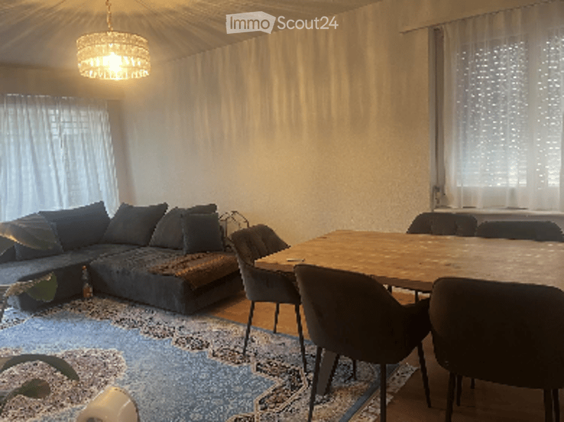Bright living and dining area with a grey sectional sofa, coffee table, dining table with chairs, and a patterned rug.
