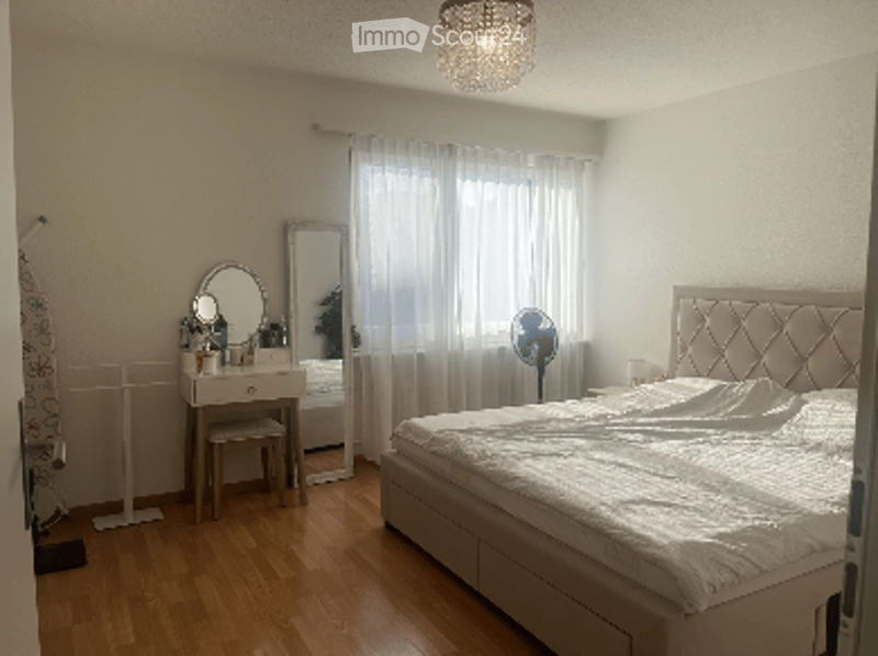 Bright bedroom with wooden flooring, large window with white curtains, a tufted headboard bed, a vanity with a mirror, and a small fan near the window.