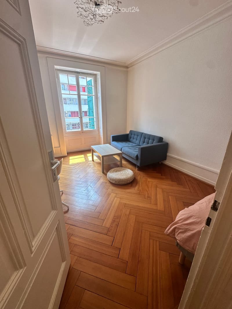 A modern living room with wooden herringbone flooring, a blue sofa, a coffee table, a round woven stool, and a bed with a pink blanket visible through an open doorway.