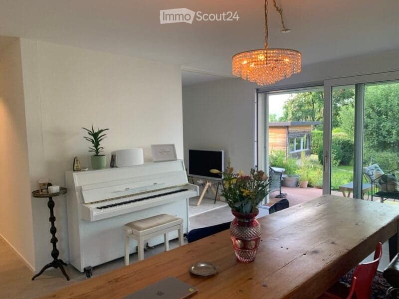 Bright living space with a white piano, wooden dining table, laptop, vase with flowers, and large windows offering a view of the garden.