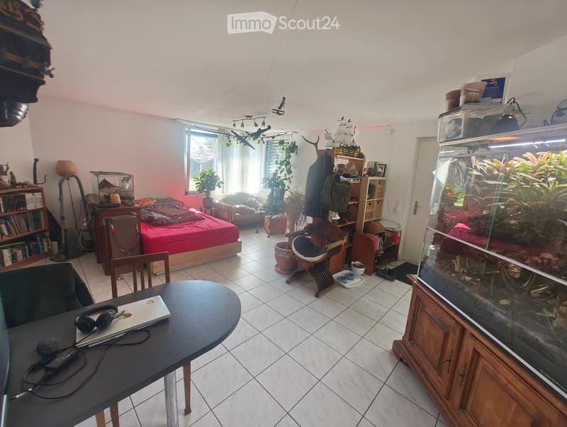 Well-lit room with a bed, sofa, fish tank, bookshelf, and a desk with a laptop and headphones. The room features tiled flooring and large windows with sheer curtains.