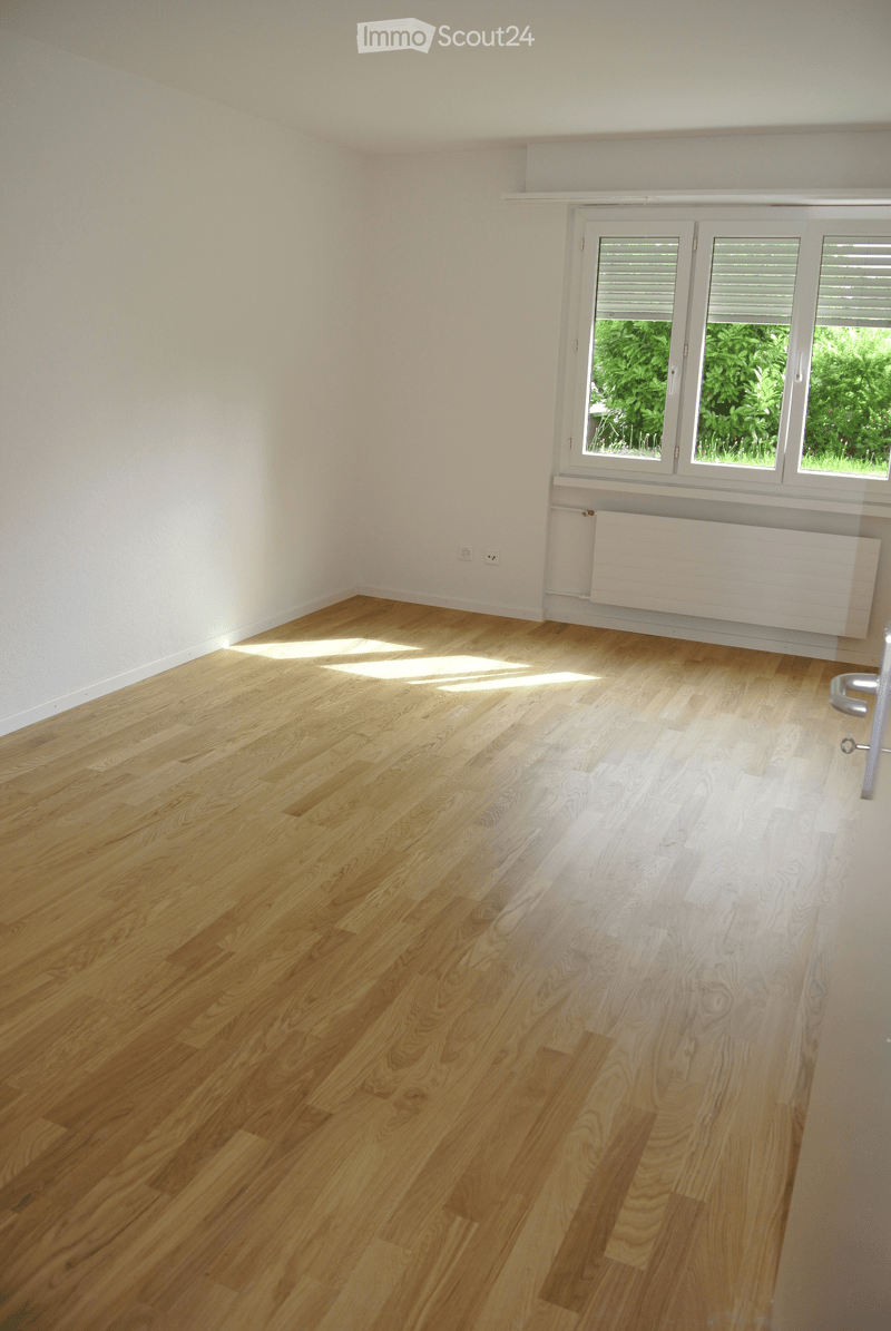 Empty room with light wooden flooring and white walls, featuring a large window with blinds and a radiator below it.