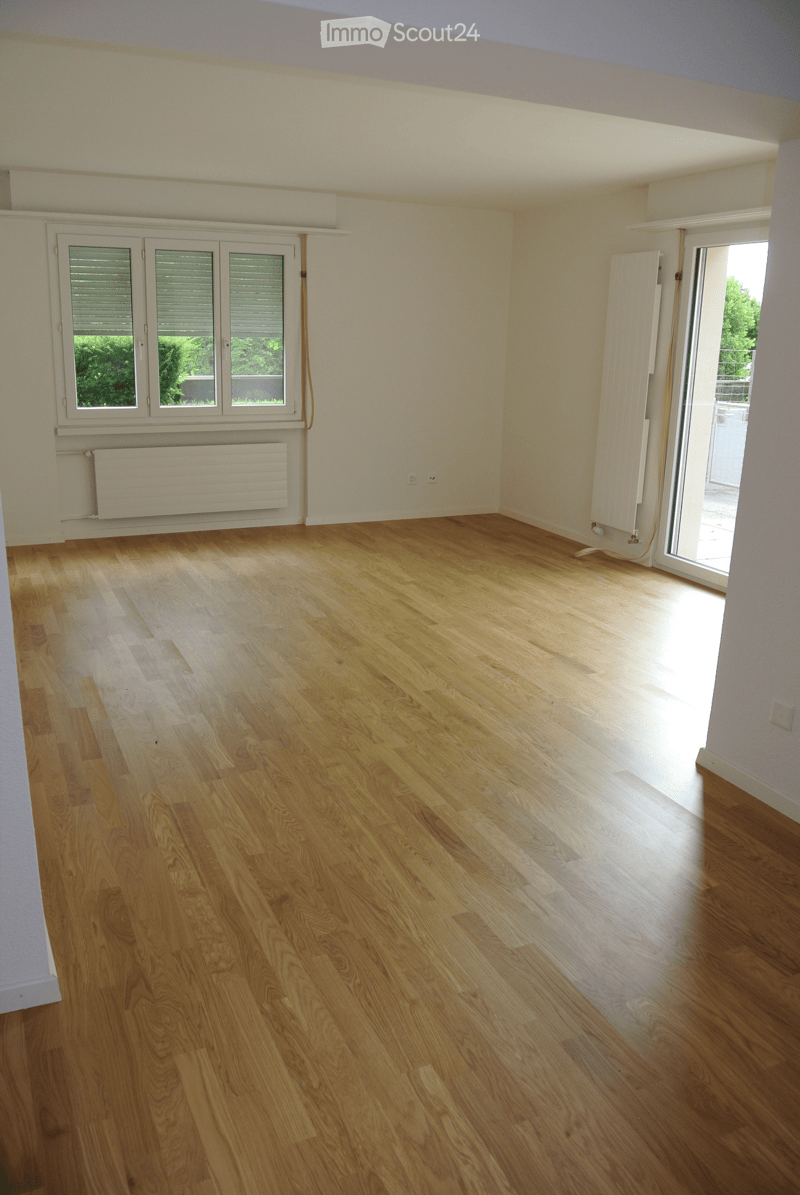 Empty room with wooden flooring, white walls, large windows, and a sliding glass door leading to a balcony.