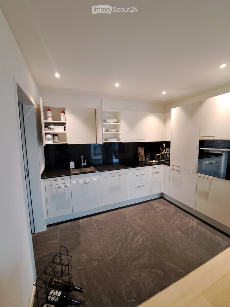 Modern kitchen with white cabinets, black countertops, stainless steel sink, and built-in oven. Includes a wine rack with several bottles, a coffee machine, and various kitchen utensils.