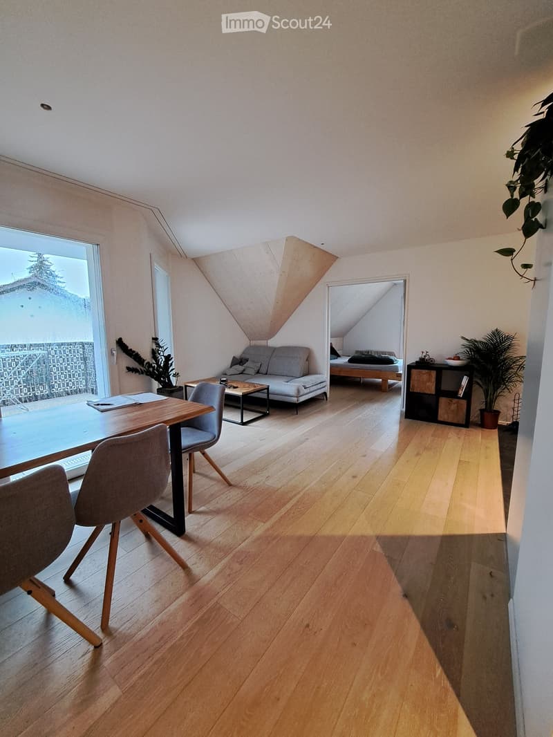 Bright, modern living and dining area with wooden flooring, large windows, a sofa, coffee table, dining table with chairs, and a small bedroom area visible through an open doorway.