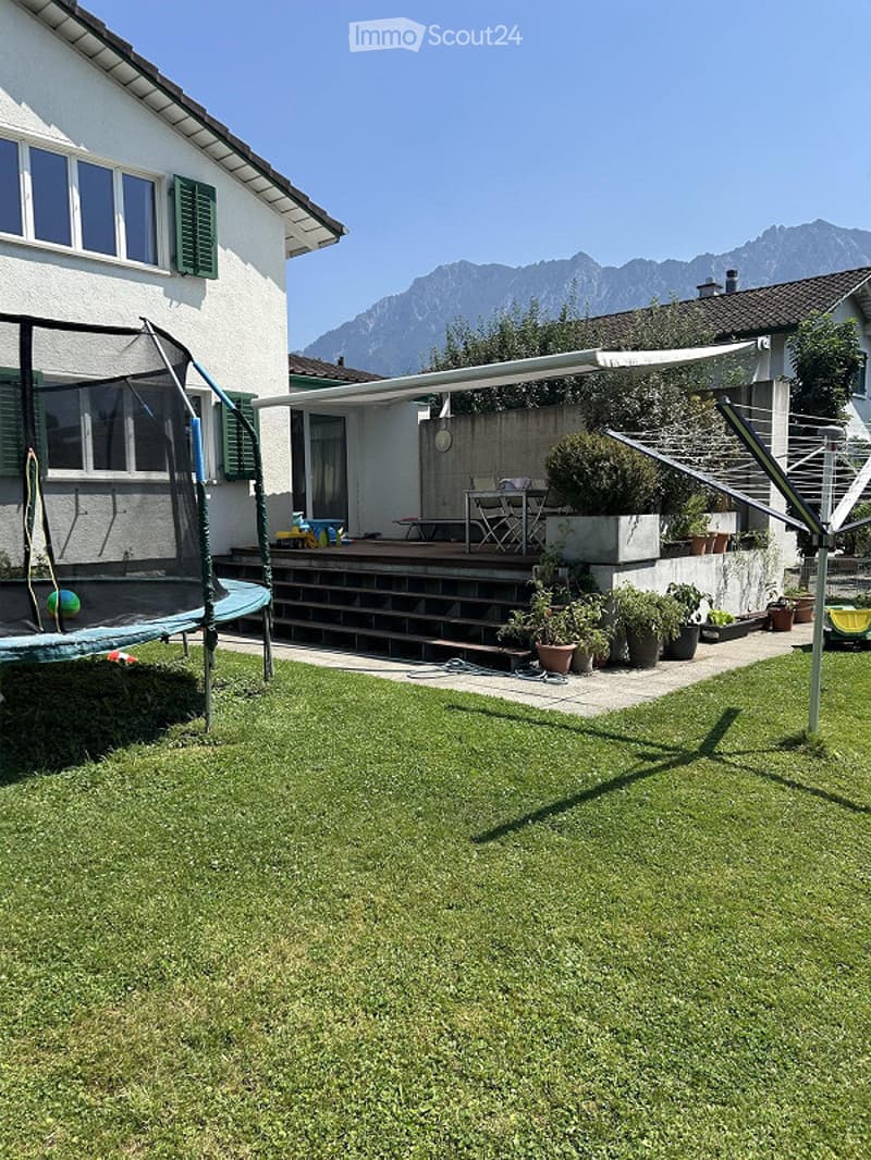 Well-maintained garden with green lawn, trampoline, patio area with table and chairs, potted plants, and scenic mountain view in the background.