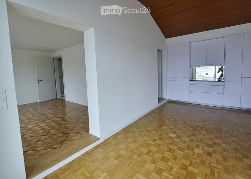 Empty room with light wooden flooring and white walls, featuring a small kitchen area with cabinets and a countertop.