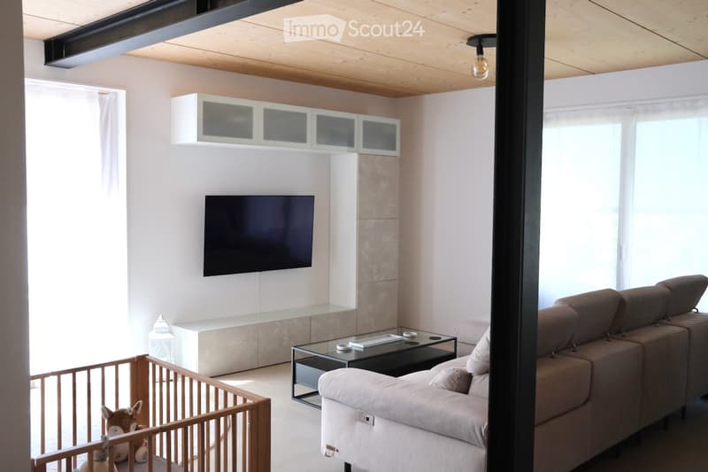 Modern living room with a grey sectional sofa, a glass coffee table, a flat-screen TV mounted on the wall, built-in white cabinets, and a small wooden playpen with a stuffed animal inside.