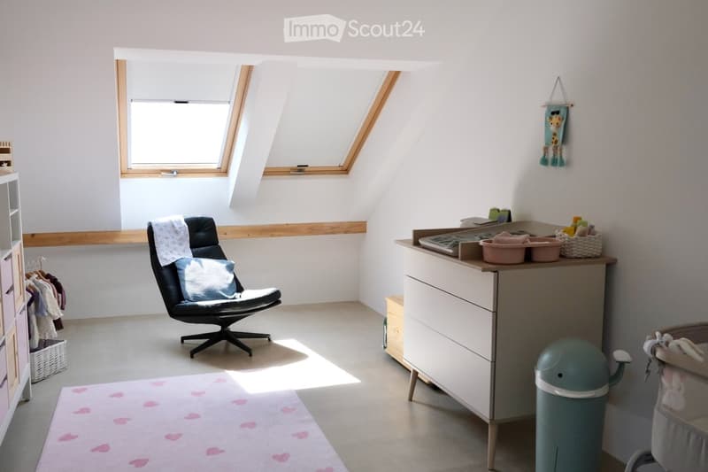 Brightly lit room with a slanted ceiling, featuring a black leather chair with a blue pillow, a baby changing table with baskets, a baby chair, and a rug with pink heart patterns.