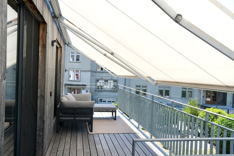 Balcony with wooden decking, modern sofa and ottoman, metal railing, and retractable awning.