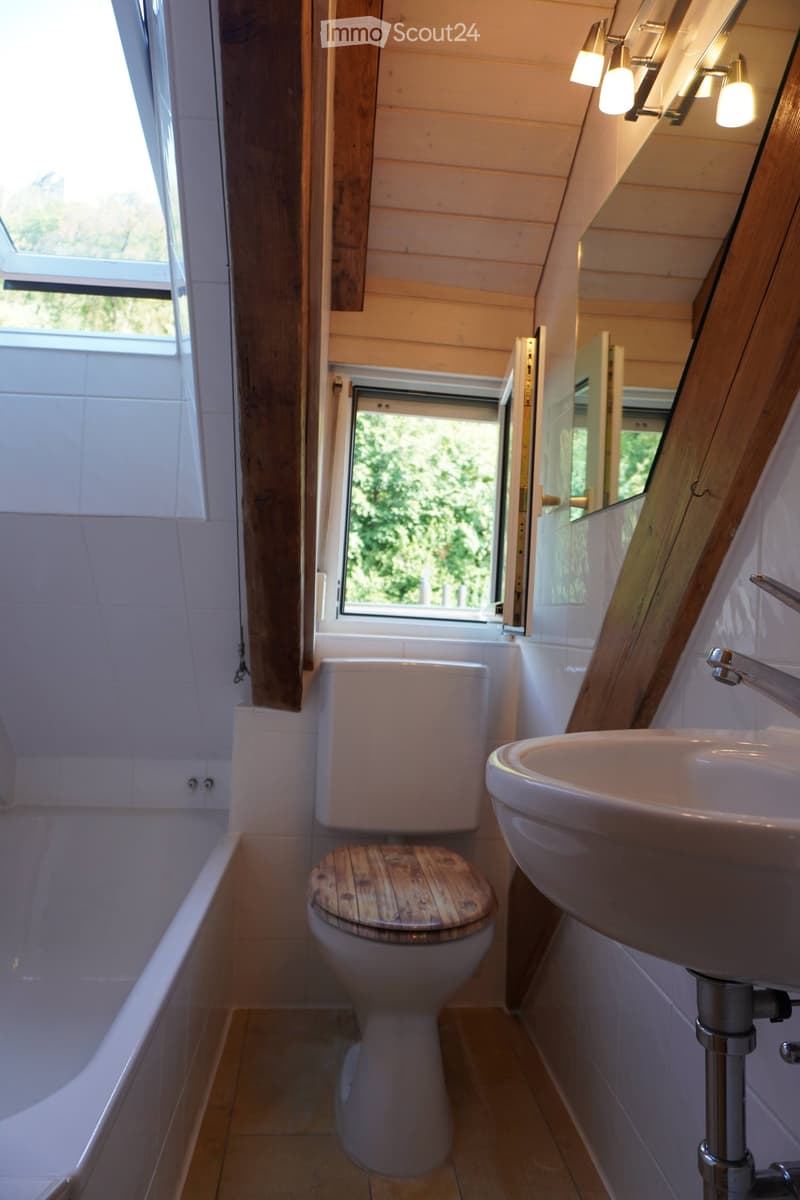 Cozy bathroom with wooden walls, a bathtub, toilet with a wooden seat, sink, and a large mirror above the sink. Natural light streams in through a window.