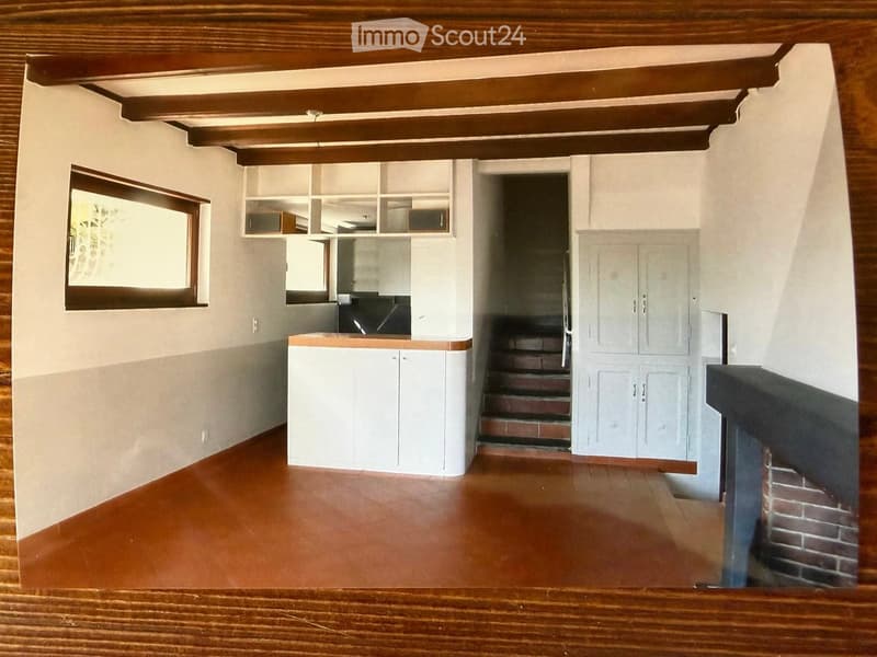 Empty room with wooden ceiling beams, white walls, and a wooden floor. Features include built-in cabinets, a window, and a staircase leading to another level.
