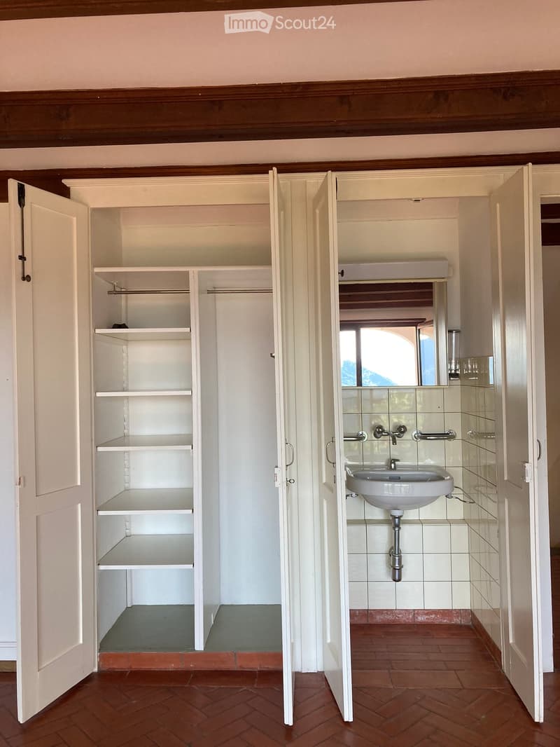 Bathroom with a white sink, mirror, and shelves in an adjacent closet. The bathroom has tiled walls and a red brick floor.