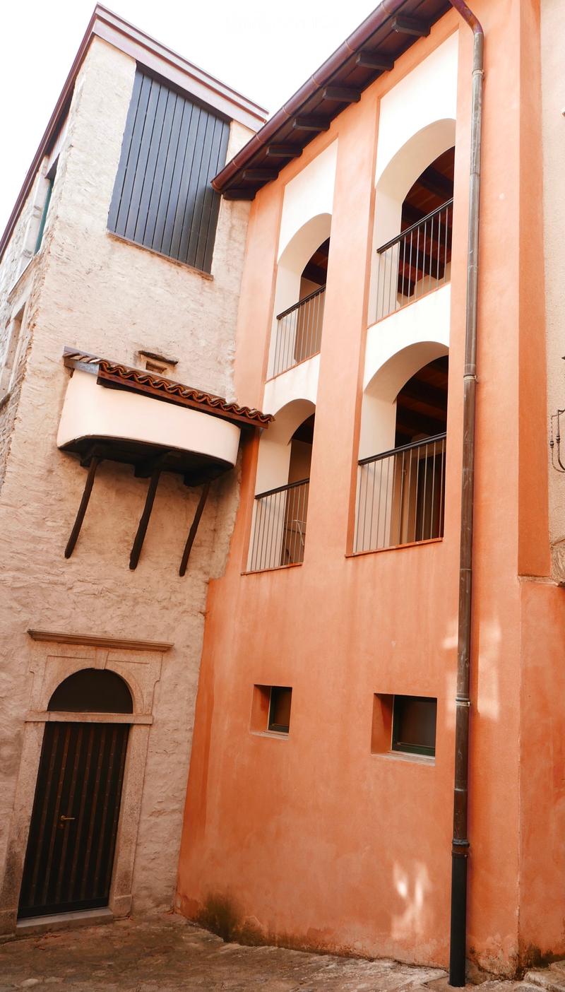 Two-story building with peach-colored walls, arched windows, and balconies. The left side features a stone facade with a small arched doorway and a protruding balcony with wooden supports. The right side has a smooth peach-colored wall with multiple arche