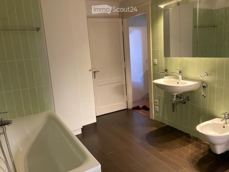 Modern bathroom with green tiled walls, dark wooden floor, bathtub, sink, toilet, and mirror. A door leads to another room.