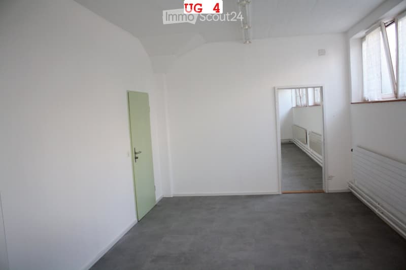 Empty room with white walls, grey carpeted floor, and a single green door. A small window with white frame is visible on the right side.