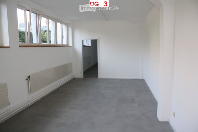 Empty room with white walls, grey carpeted floor, large windows, radiator, and an open doorway leading to another space.