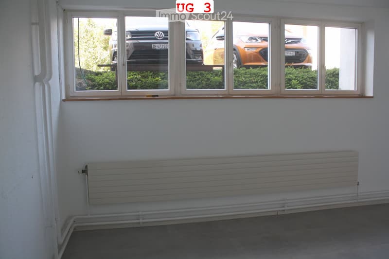 Empty room with white walls, large windows showing parked cars and greenery, radiator, and a grey floor.
