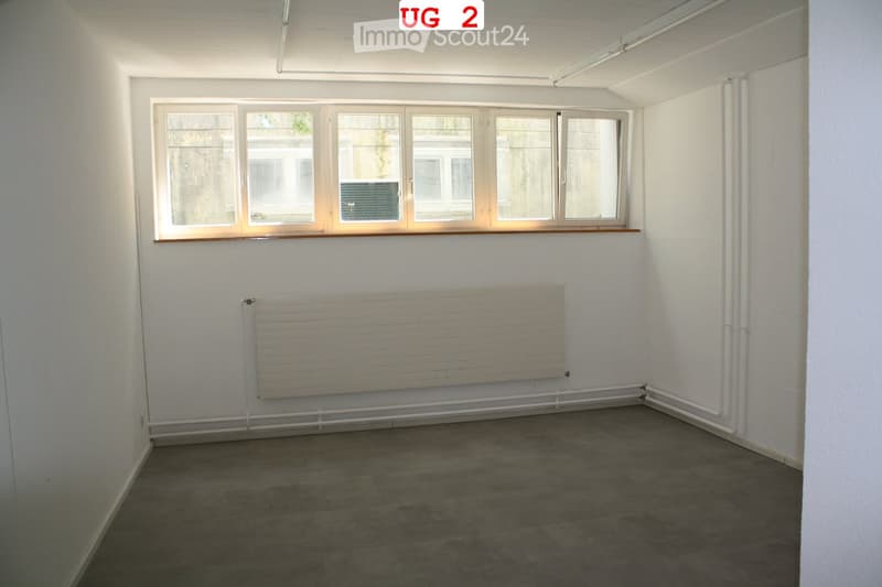 Empty room with white walls, grey tiled floor, large window with wooden frame, radiator, and visible pipes along the wall.
