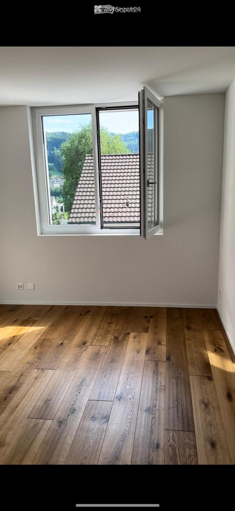 Empty room with wooden flooring and large windows offering a view of the roof and surrounding greenery.