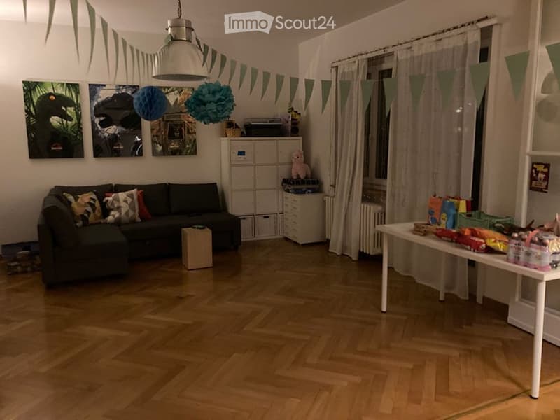 A cozy living room with a dark green sectional sofa, colorful throw pillows, a white coffee table, a storage unit with a pink stuffed animal, and a table filled with children's toys and decorations.