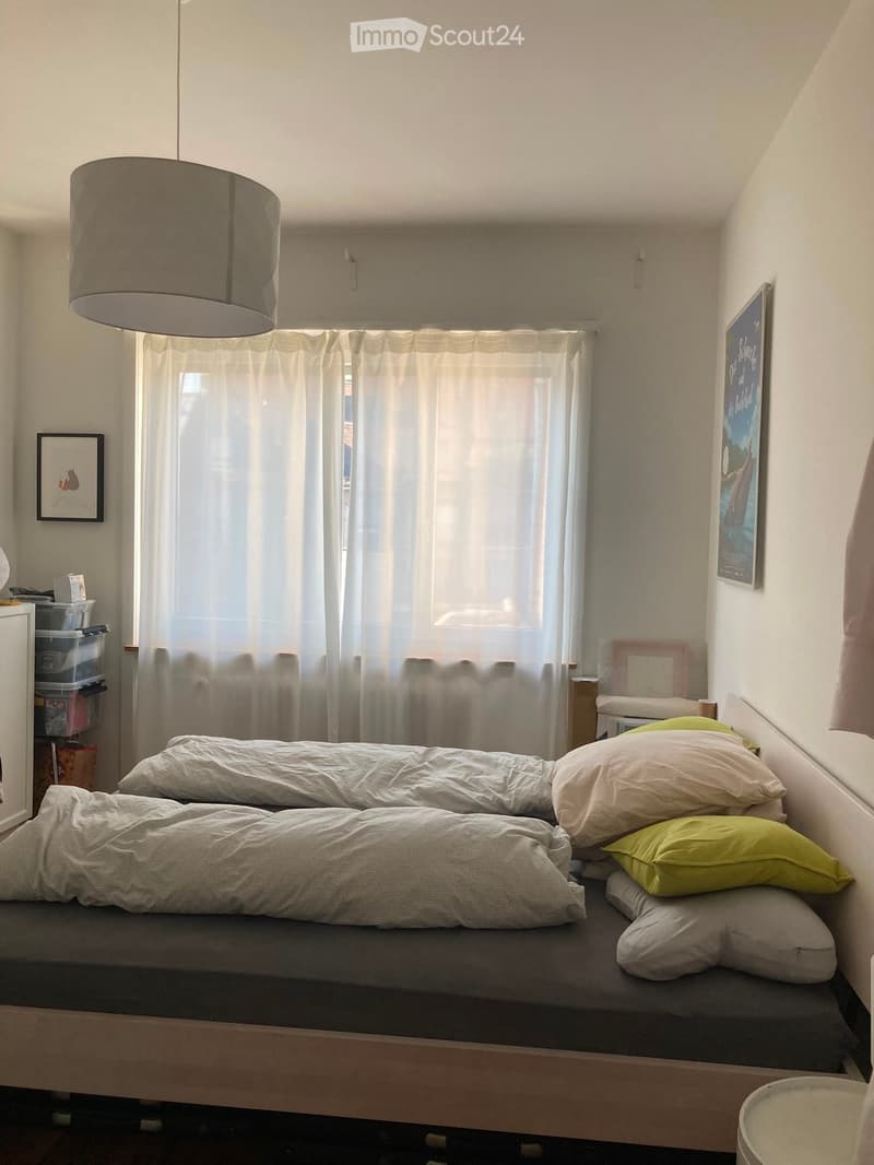 Cozy bedroom with a single bed, multiple pillows, a window with white curtains, a hanging lamp, and a small storage unit.