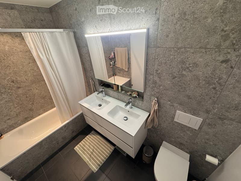 Modern bathroom with a double sink, large mirror, bathtub, and toilet. Features marble walls and a striped bath mat.