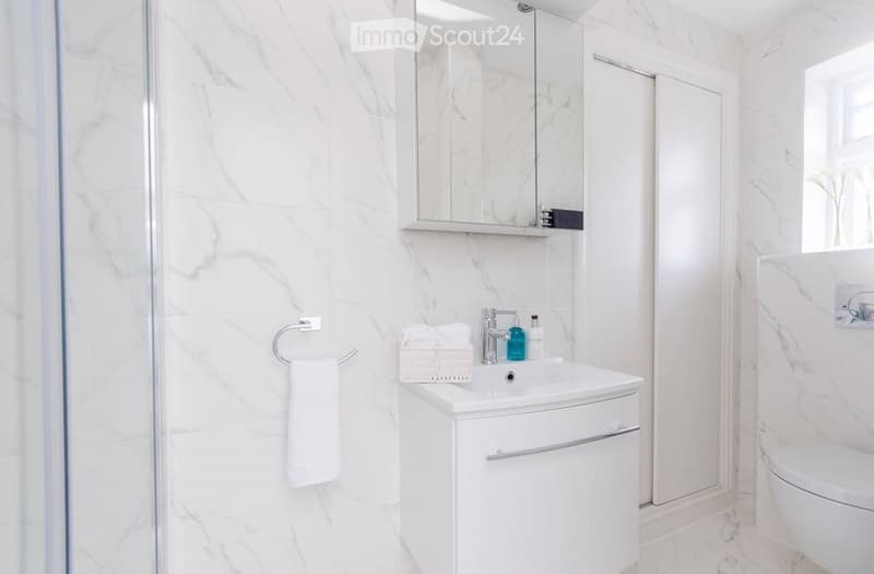 Modern bathroom with white marble walls and floor, a white sink with a cabinet, a mirror above the sink, a towel on a ring, and a toilet in the corner.