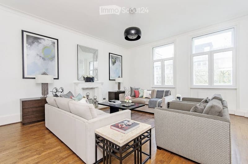 Bright living room with white walls, wooden flooring, and large windows. Features a white sofa, grey armchair, coffee table, side table, fireplace, and decorative items.