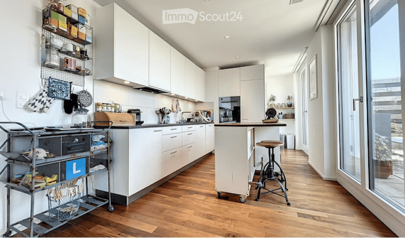 Modern kitchen with white cabinets, wooden flooring, and various appliances including a sink, stove, and microwave. A small island with a stool is present, along with a cart holding various items.