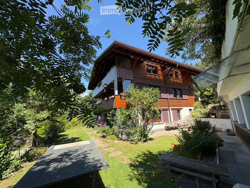 A well-maintained garden area with a wooden bench, a small table, and a cat visible. The garden is surrounded by lush greenery and leads to a two-story wooden house with a balcony and multiple windows.