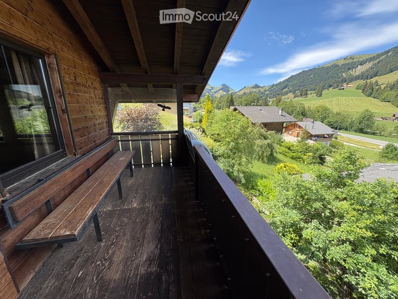 Wooden balcony with a bench, overlooking a scenic mountain landscape with green fields and scattered houses.