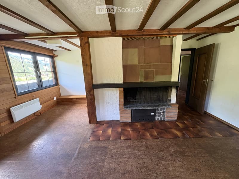 Empty room with wooden beams on the ceiling, a fireplace with a tiled surround, wooden flooring, and large windows offering a view of greenery.
