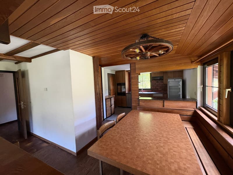 A cozy dining area with a wooden table and chairs, connected to a kitchen with wooden cabinets, a refrigerator, and an oven. The room features wooden ceilings and large windows.