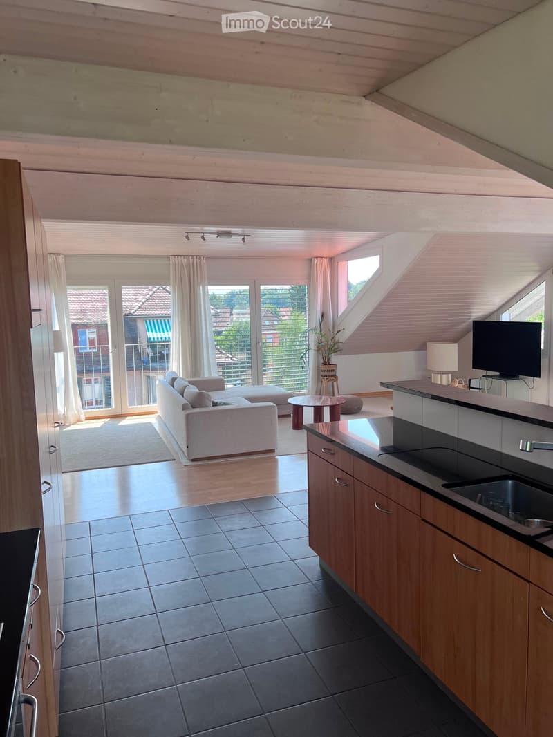 Open-plan living and kitchen area with wooden beams, large windows, and a balcony. Features a sofa, coffee table, television, and modern kitchen with cabinets and a sink.