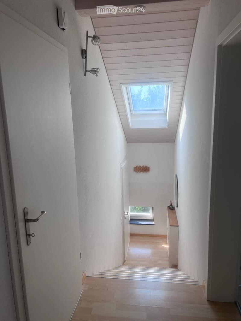 Modern hallway with wooden ceiling, white walls, and a skylight window at the top. Features a small wooden shelf with decorative items and a small table with a lamp.