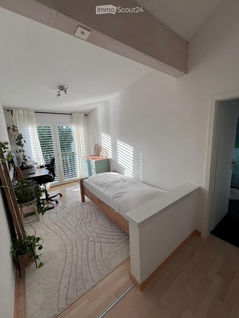 Bright bedroom with a wooden bed, desk, chair, laptop, and a plant near the window. The room features white walls, wooden flooring, and large windows with blinds.