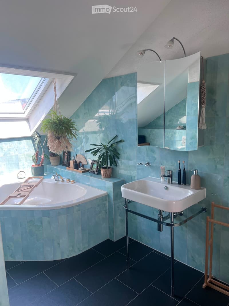 Modern bathroom with blue tiles, a bathtub, a sink, a large mirror, and decorative plants on the windowsill.