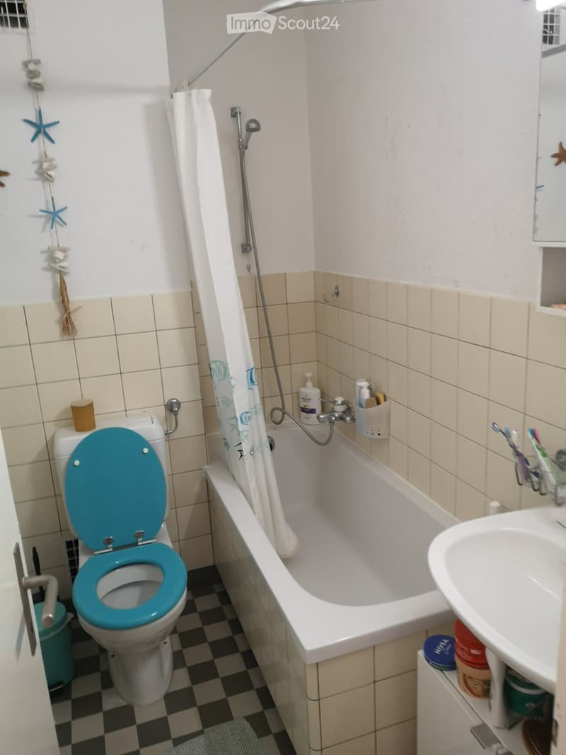 Bathroom with a white bathtub, a toilet with a turquoise seat cover, a sink, and a toothbrush holder. The walls are tiled in beige and white, and the floor has a black and white checkered pattern.