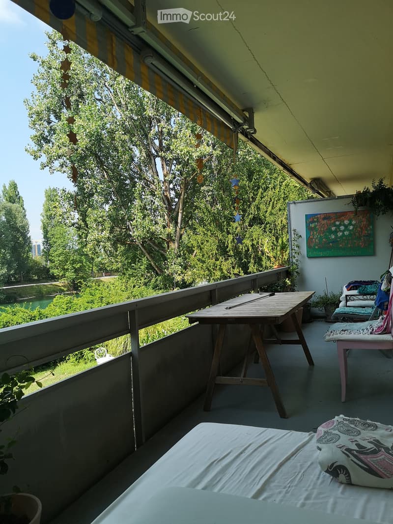 Balcony with a wooden table, chairs, a bed with white sheets, and a colorful painting on the wall. The balcony has a view of greenery and a river.