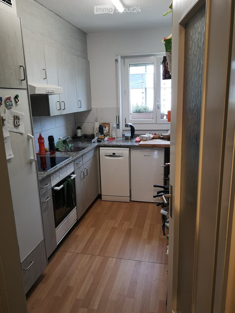 Modern kitchen with light wooden flooring, white cabinets, stainless steel appliances including a refrigerator, oven, and dishwasher. Countertop has various items including a coffee maker, cutting board, and bowls.