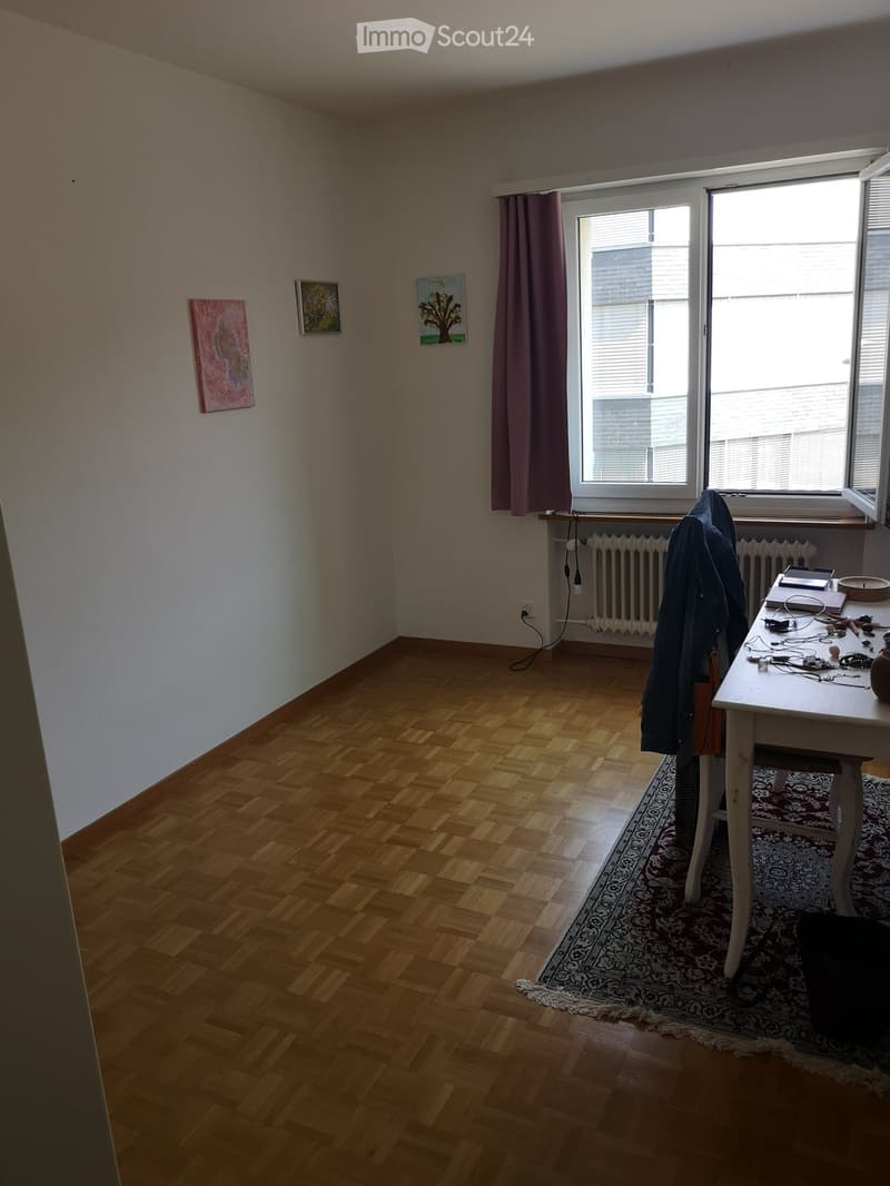 A small, unfurnished room with wooden flooring, a large window with curtains, a radiator, and a small table with a laptop and other items.