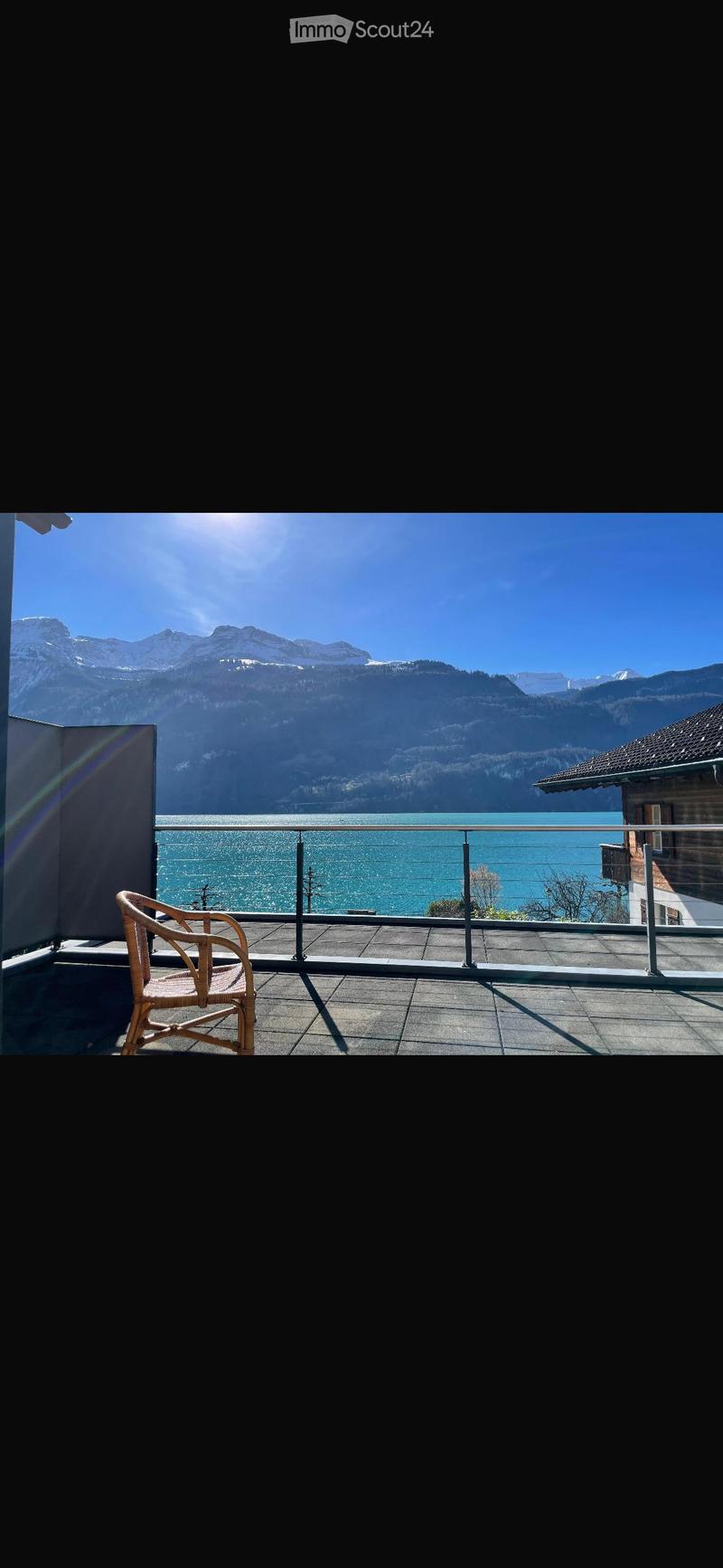 Balcony with a view of a lake and mountains, featuring a single wicker chair and a metal railing.