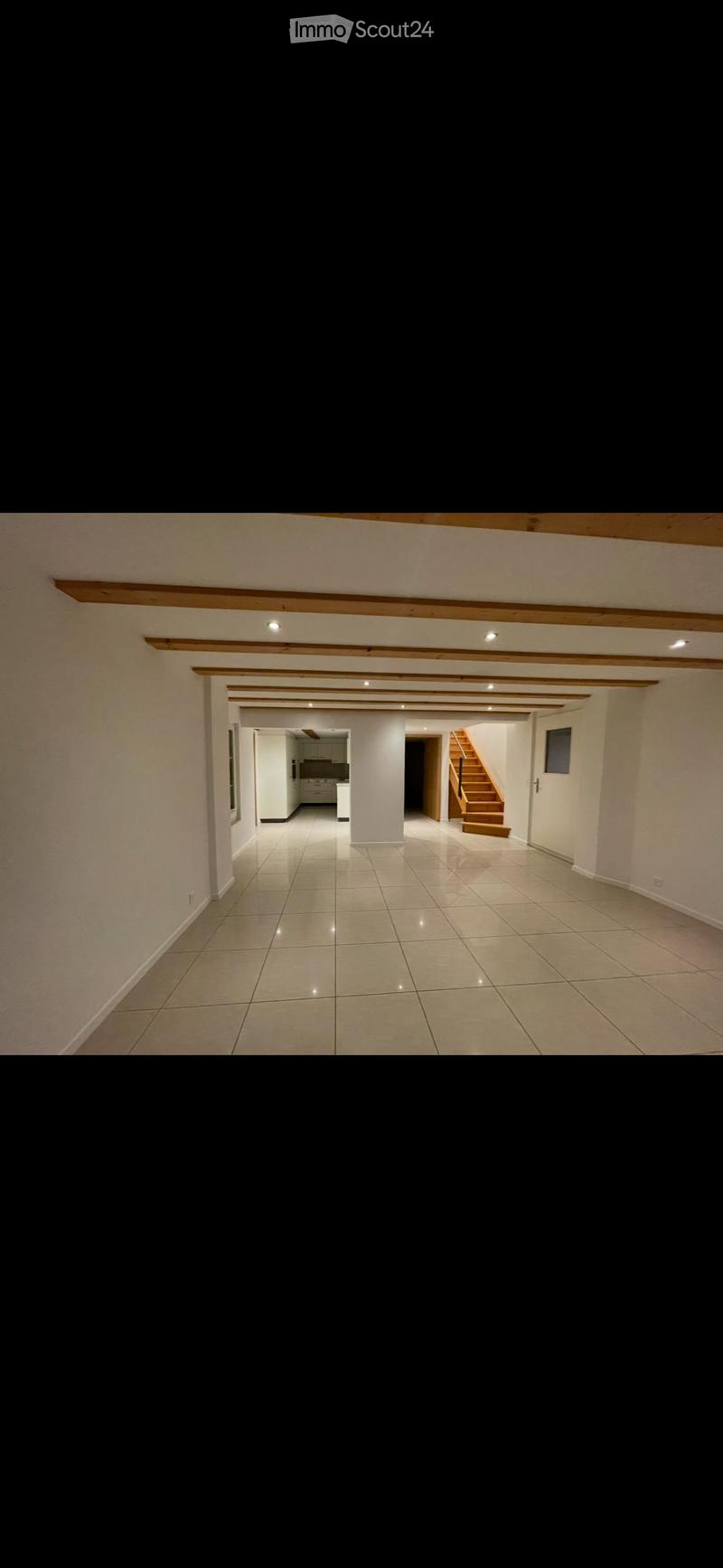 Empty hallway with tiled floor, white walls, wooden beams on the ceiling, and visible rooms including a kitchen and staircase in the background.
